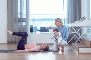Äiti treenaa keskivartalon lihaksia lattialla ja personal trainer kannustaa pitäen vauvaa sylissä,