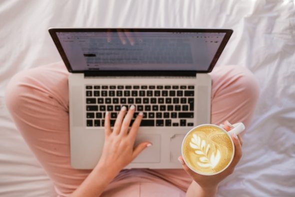 Nainen istuu sängyllä sylissään kannettava tietokone ja kädessä kahvikuppi. Jalassa on vaalenpunaiset housut. Kuva on otettu yläkulmasta.