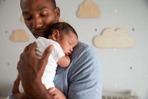 Isä pitää nukkuvaa, vastasyntynyttä vauvaa rintaansa vasten. Lastenhuoneen seinällä on pilvenmuotoisia koristeita.