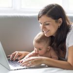 Äiti makoilee vauvan kanssa sohvalla ja katselee hymyillen tietokonetta.