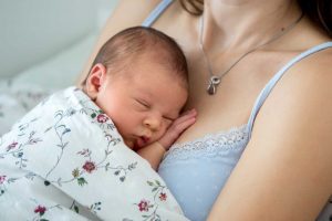 Pieni vauva nukkuu äidin rintakehää vasten. Äidillä on päällään sininen toppi ja vauvalla vaalea kukallinen peitto.