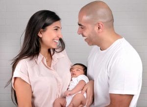 Vaaleisiin vaatteisiin pukeutuneet äiti ja isä katsovat toisiinsa iloisina. Äiti pitää sylissään pientä, tummatukkaista vauvaa.