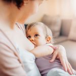 Taaperoikäinen lapsi on äidin rinnalla ja pitää toista kättään äidin vapaan rinnan päällä, katsoen samalla äitiä tiukasti silmiin.