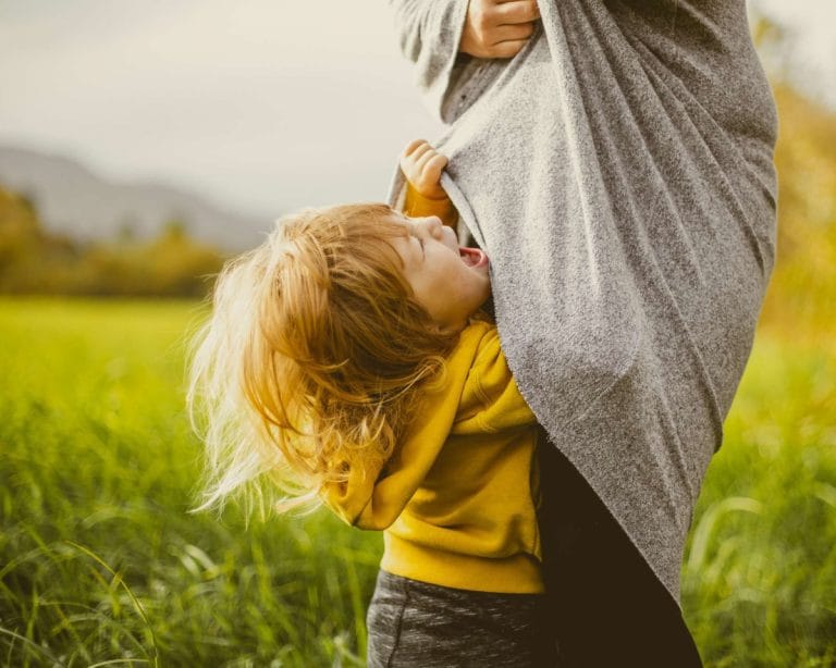 Taaperoikäinen lapsi itkee ja roikkuu äidin paidassa ulkona.