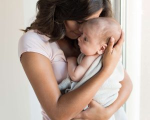 Tummahiuksinen äiti halaa pientä, peittoon käärittyä vauvaa sylissään.