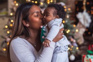 Äiti nostaa hymyilevää, pientä vauvaa kainaloista ja suukottaa tätä poskelle. Taustalla näkyy joulukuusen valot ja vaaleat koristeet.