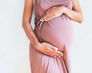 Vaalenpunaiseen mekkoon pukeutunut odottava nainen pitää käsiään suuren raskausvatsan ympärillä vaaleaa taustaa vasten.
