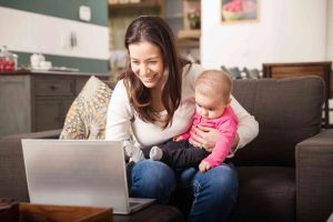 Nuori tummahiuksinen nainen istuu olohuoneen sohvalla sylissään vaaleanpunaiseen paitaan puettu puolivuotias vauva. Nainen katsoo iloisena kannettavan tietokoneen ruutua.