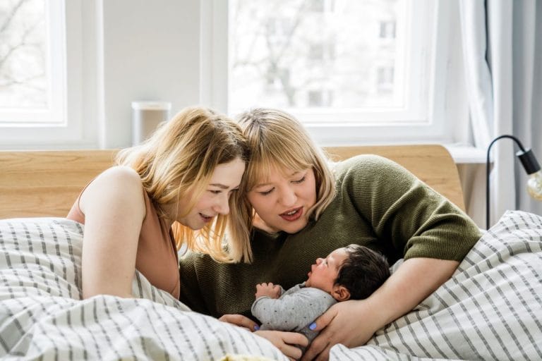 Vauvan kaksi äitiä ihailevat vauvaa aamulla sängyssä.