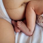 Lähikuva vastasyntyneen imetystilanteesta. Kuvassa näkyy vanhemman mahaa ja verkkohousut sekä vauvan jalat.