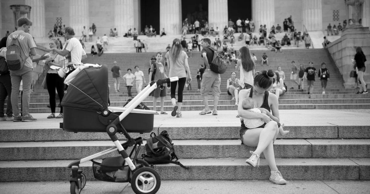 Vanhempi istuu vilkkaassa turistikohteessa kiviportailla ja imettää vauvaa.