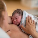 Pyyheliinalla peitelty, kosteahiuksinen vastasyntynyt katsoo äitiä ihokontaktissa